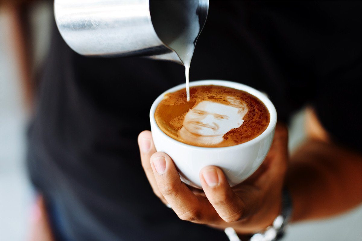 A barista is pouring steamed milk into a cup of espresso to create latte art with a face, with a hand holding the cup and a blurred dark background.